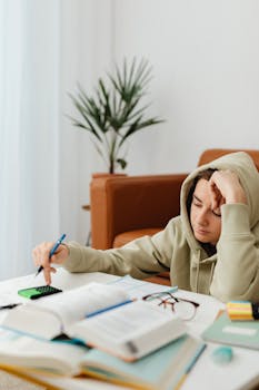 Teenage boy in a hoodie studying at home, looking tired, with books and a calculator.
